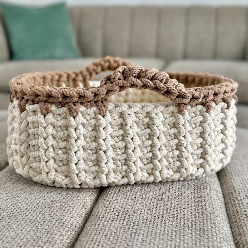 May include: A round, hand-crocheted basket featuring a thick, cream-colored base with a vertical ribbed texture and a contrasting brown rim. The basket includes two integrated, braided handles on opposite sides, crafted from the same brown yarn.