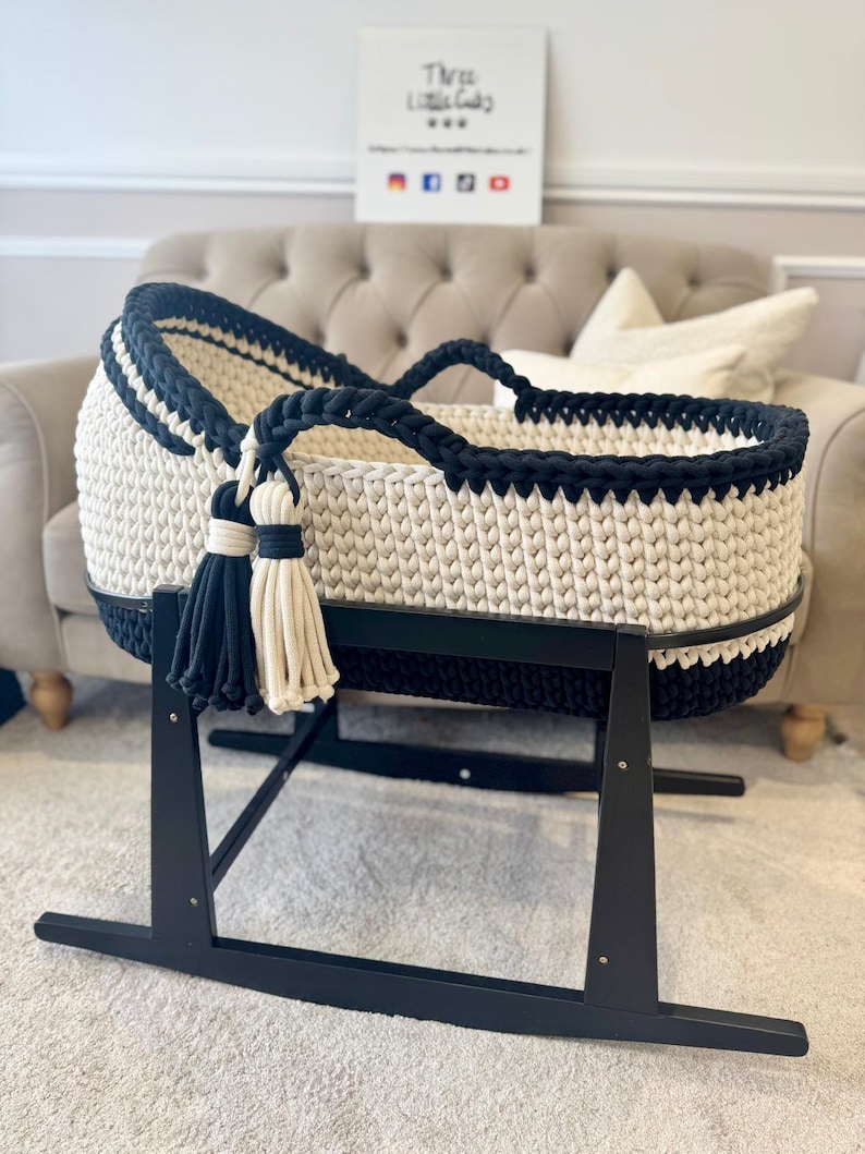 May include: A handmade, cream-colored crochet baby bassinet with navy blue trim and handles. The bassinet sits on a black wooden rocking stand and features decorative tassels. The text "Three Little Gals" is visible in the background.