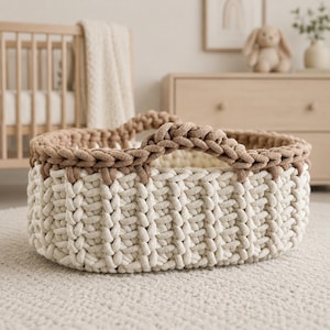 May include: A round, hand-crocheted storage basket featuring a thick, cream-colored base and a contrasting tan-colored rim with two integrated handles. The basket is constructed from chunky, braided yarn, creating a textured, ribbed pattern throughout.