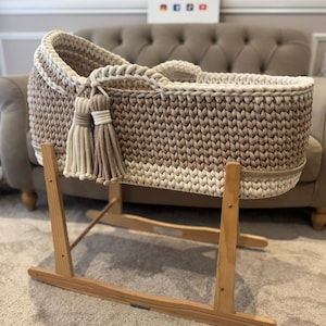 May include: A handmade, neutral-toned baby bassinet with a woven design. The bassinet features a light brown and cream color scheme, with decorative tassels. It is mounted on a wooden rocking stand.