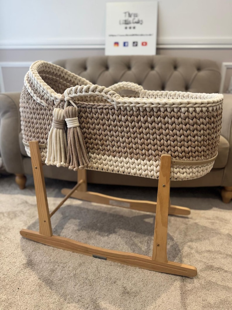May include: A handmade, neutral-toned baby bassinet with a woven design. The bassinet features a light brown and cream color scheme, with decorative tassels. It is mounted on a wooden rocking stand.