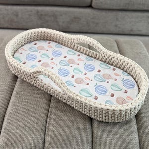 Puede incluir: Una cesta para cambiar pañales ovalada de color crema con un diseño tejido. El interior presenta una tela estampada con globos aerostáticos de acuarela en azul, verde y marrón. La cesta está colocada sobre una superficie tapizada gris.
