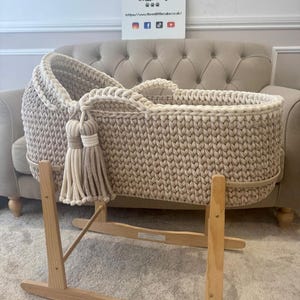 May include: A beige and cream-colored crochet baby bassinet with a wooden rocking stand. The bassinet features a thick, textured weave and decorative tassels. The sign in the background reads "Three Little Cubs".