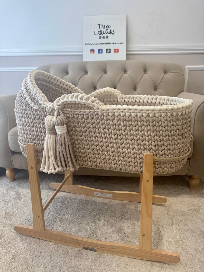 May include: A beige and cream-colored crochet baby bassinet with a wooden rocking stand. The bassinet features a thick, textured weave and decorative tassels. The sign in the background reads "Three Little Cubs".