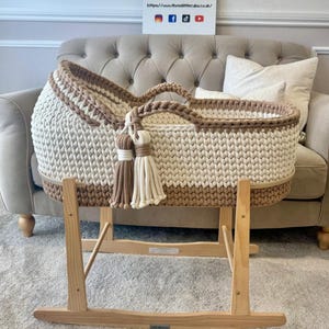 May include: A handmade, cream and tan crochet baby bassinet with a wooden rocking stand. The bassinet features a woven design, a tan trim, and decorative tassels. The wooden stand is light-colored and has a rocking base. The sign in the background says "Three Little Cubs".