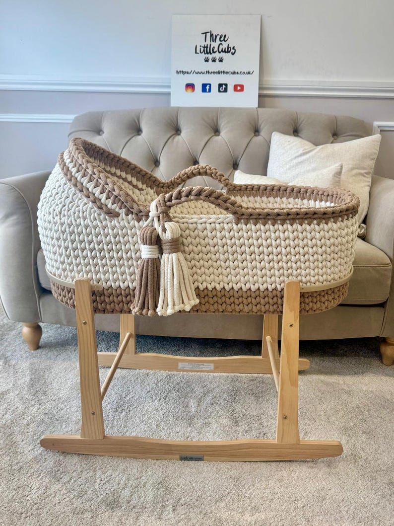 May include: A handmade, cream and tan crochet baby bassinet with a wooden rocking stand. The bassinet features a woven design, a tan trim, and decorative tassels. The wooden stand is light-colored and has a rocking base. The sign in the background says "Three Little Cubs".