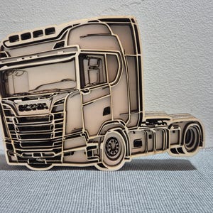 Puede incluir: Una pieza de arte mural de madera en capas con forma de camión Scania. El diseño presenta madera de color claro con contornos oscuros cortados con láser que definen la cabina, la parrilla, las ruedas y el chasis del camión en relieve.