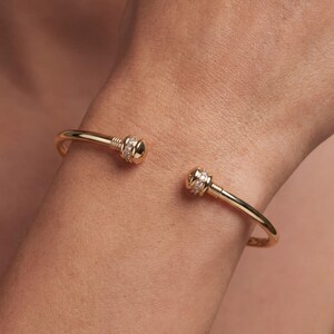 Op de afbeelding: Een goudkleurige open armband met een gladde, gepolijste band. Elk uiteinde van de armband eindigt in een afgeronde, koepelvormige dop, geaccentueerd met een band van kleine, heldere, fonkelende steentjes in het metalen oppervlak.