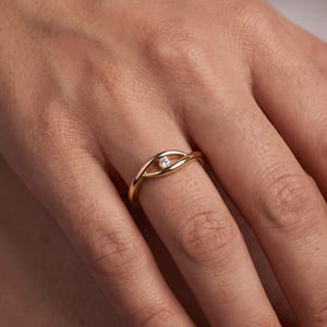 Könnte beinhalten: Ein goldener Ring mit einer glatten, polierten Ringschiene, die sich oben in zwei geschwungene Linien teilt und eine augenähnliche Form bildet. Ein einzelner, kleiner, klarer, rund geschliffener Edelstein ist mittig in dieser Öffnung gefasst.