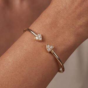 Puede incluir: Una pulsera abierta de tono dorado con dos terminales triangulares, cada uno engastado con tres pequeñas piedras claras y brillantes. La banda de metal tiene un acabado liso y pulido con un perfil delgado, creando una apariencia refinada.