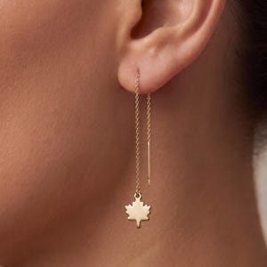 Pode incluir: Um brinco de fio dourado com uma corrente delicada que passa pelo lóbulo da orelha e termina num pequeno pendente plano em forma de folha de ácer. O design inclui uma haste metálica fina e reta no topo da corrente para fácil inserção.