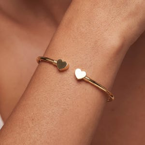 May include: A gold-toned open cuff bracelet featuring two small, polished heart-shaped charms at each end. The band has a smooth, rounded profile with subtle ribbed detailing where the hearts connect to the main wire structure of the bracelet.