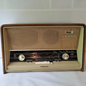 Op de afbeelding: Vintage Philips radio met een houten frame, beige voorpaneel en een geweven luidsprekerkap. Voorzien van twee grote afstemknoppen, een frequentieschaal en diverse witte knoppen. De radio heeft een klassiek mid-century modern design.
