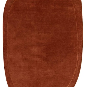 May include: An oval-shaped rug in a solid, deep terracotta orange color. The rug features a smooth, uniform pile texture throughout its surface and a distinct, slightly raised border that follows the rounded, organic contour of the rug's perimeter.