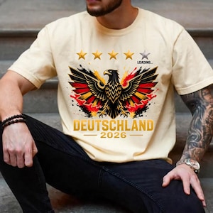 Op de afbeelding: Een crèmekleurig t-shirt met een afbeelding van een zwarte adelaar met rode en gele vleugelaccenten. Boven de adelaar staan vier gouden sterren en één grijze ster met de tekst LOADING... eronder. Het woord DEUTSCHLAND en 2026 staan onderaan.