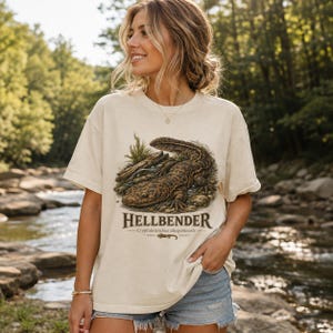 May include: A cream-coloured short-sleeve t-shirt featuring a detailed brown illustration of a hellbender salamander on a rocky riverbed. The text HELLBENDER and Cryptobranchus alleganiensis is printed below the graphic with a small salamander icon.
