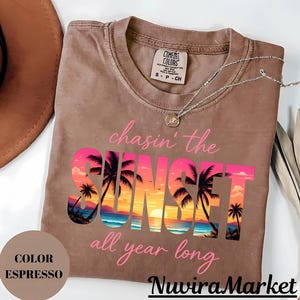 May include: An espresso-colored t-shirt featuring the text 'chasin' the SUNSET all year long' in pink script and block lettering. The word 'SUNSET' contains a graphic of a beach scene with palm trees and a sunset. A silver necklace rests on the shirt.