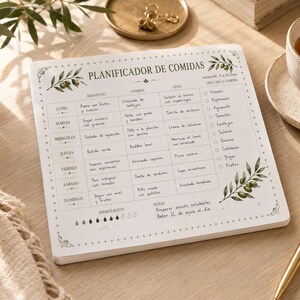 Puede incluir: Un planificador de comidas blanco con un diseño de rama de olivo impreso y el texto "PLANIFICADOR DE COMIDAS". El planificador tiene secciones para el desayuno, el almuerzo, la cena y una lista de compras. Un bolígrafo dorado y una taza de té también son visibles.