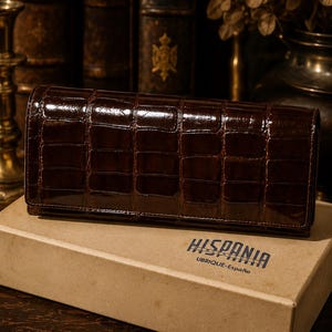 May include: A dark brown leather wallet with a crocodile skin texture, displayed on a cardboard box. The wallet is rectangular and shiny. The box has the word "HISPANIA" printed on it.
