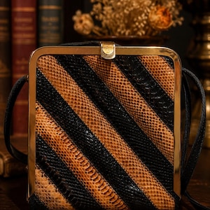 May include: A square, vintage handbag with a gold-toned frame and clasp. The bag features a diagonal striped pattern in black and tan, with a black shoulder strap. The bag is likely made of leather or a similar material.