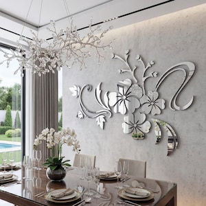 May include: A dining room scene featuring a large, reflective floral wall art installation. The artwork is silver and reflects the room's details. A crystal chandelier hangs above a long dining table set with plates, glasses, and silverware. A vase of white flowers is on the table.