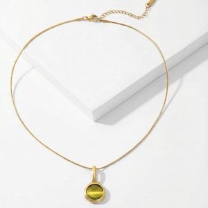 Op de afbeelding: Een goudkleurige ketting met een fijne ketting en een hanger. De hanger heeft een ronde, olijfgroene steen in een gouden bezel. De ketting wordt getoond op een wit oppervlak.