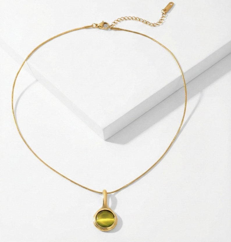Op de afbeelding: Een goudkleurige ketting met een fijne ketting en een hanger. De hanger heeft een ronde, olijfgroene steen in een gouden bezel. De ketting wordt getoond op een wit oppervlak.