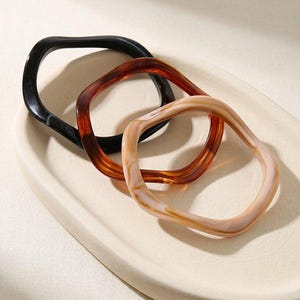 Op de afbeelding: Drie harsarmbanden in zwart, bruin en crème. De vierkante armbanden hebben een golvend design en worden gepresenteerd op een crèmekleurig, ovaal dienblad. De armbanden zijn een stijlvol accessoire.