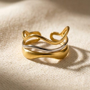 Puede incluir: Un anillo dorado con un diseño único y fluido. El anillo presenta tres bandas: dos doradas y una plateada. La banda superior tiene un diseño abierto y ondulado. El anillo se muestra sobre una superficie texturizada de color blanquecino.