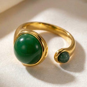 Peut inclure: Bague dorée ornée d'une grosse pierre verte ronde. La bague a un design ouvert avec une petite pierre verte à l'extrémité opposée. Fond blanc.