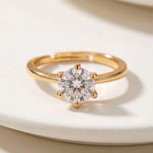 Peut inclure: Une bague dorée avec une grosse pierre précieuse ronde et transparente. La pierre est sertie de six griffes. La bague est réglable. La bague est sur une surface blanche. Bijoux.