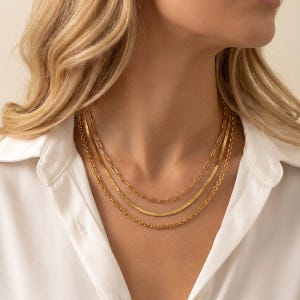 May include: A set of layered gold necklaces. The chains have various designs, including herringbone, paperclip, and rope styles. The jewellery is shown against a white shirt.