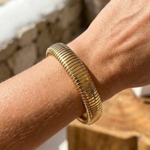 Puede incluir: Una pulsera dorada con un diseño acanalado. La pulsera es gruesa y se envuelve alrededor de la muñeca. La pulsera está hecha de metal y tiene un acabado brillante. La pulsera es una joya.