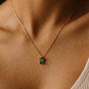 Puede incluir: Un collar de cadena dorada con un colgante de piedra preciosa verde de corte esmeralda rectangular. El colgante está sujeto por un engaste sencillo de cuatro garras y cuelga de una delicada cadena de eslabones dorados sobre la clavícula.