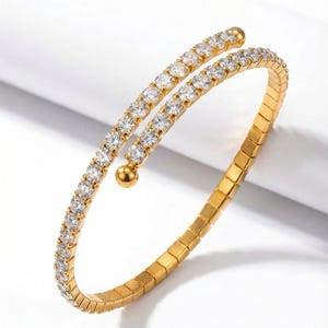 May include: A gold-toned bracelet with a spiral design. The bracelet features a double band, one adorned with clear, sparkling stones. The other band is a solid gold color. A small gold ball accents the design. The bracelet is on a white background.