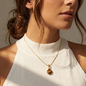 Op de afbeelding: Een goudkleurige ketting met een hanger met een ronde, bruine tijgeroogsteen. De hanger hangt aan een fijne ketting. De ketting wordt gedragen over een witte top.