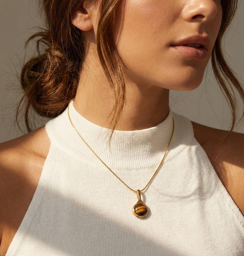 Op de afbeelding: Een goudkleurige ketting met een hanger met een ronde, bruine tijgeroogsteen. De hanger hangt aan een fijne ketting. De ketting wordt gedragen over een witte top.