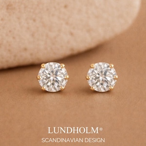 Peut inclure: Une paire de boucles d'oreilles à clous avec des pierres rondes à facettes transparentes serties dans des griffes en métal doré. Le texte LUNDHOLM SCANDINAVIAN DESIGN est imprimé en bas. Les boucles d'oreilles ont une forme circulaire classique.