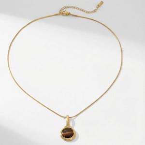 Op de afbeelding: Een goudkleurige ketting met een hanger. De hanger heeft een ronde, bruine steen in een gouden bezel. De ketting hangt aan een fijne gouden ketting. Gepresenteerd tegen een witte achtergrond.