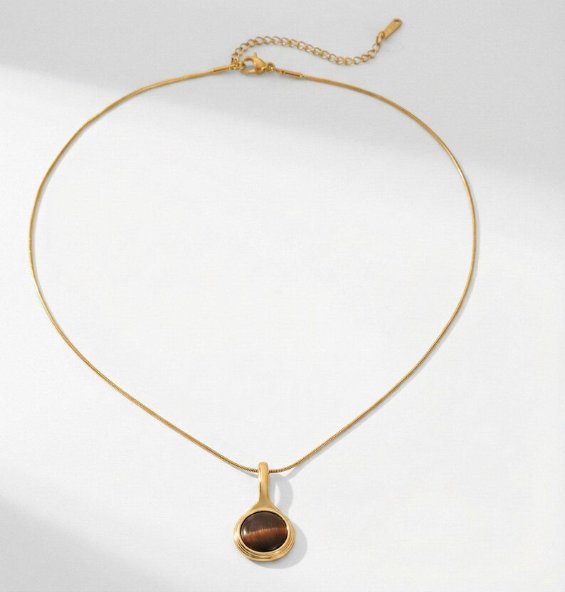 Op de afbeelding: Een goudkleurige ketting met een hanger. De hanger heeft een ronde, bruine steen in een gouden bezel. De ketting hangt aan een fijne gouden ketting. Gepresenteerd tegen een witte achtergrond.