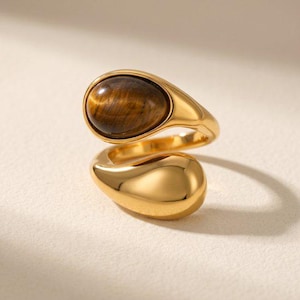 Op de afbeelding: Een goudkleurige open ring met een gepolijste, druppelvormige band. Het ene uiteinde van de ring is bezet met een gladde, ovaalvormige bruine tijgeroog-edelsteen, terwijl het andere uiteinde een massieve, gladde goudkleurige metalen druppelvorm heeft.