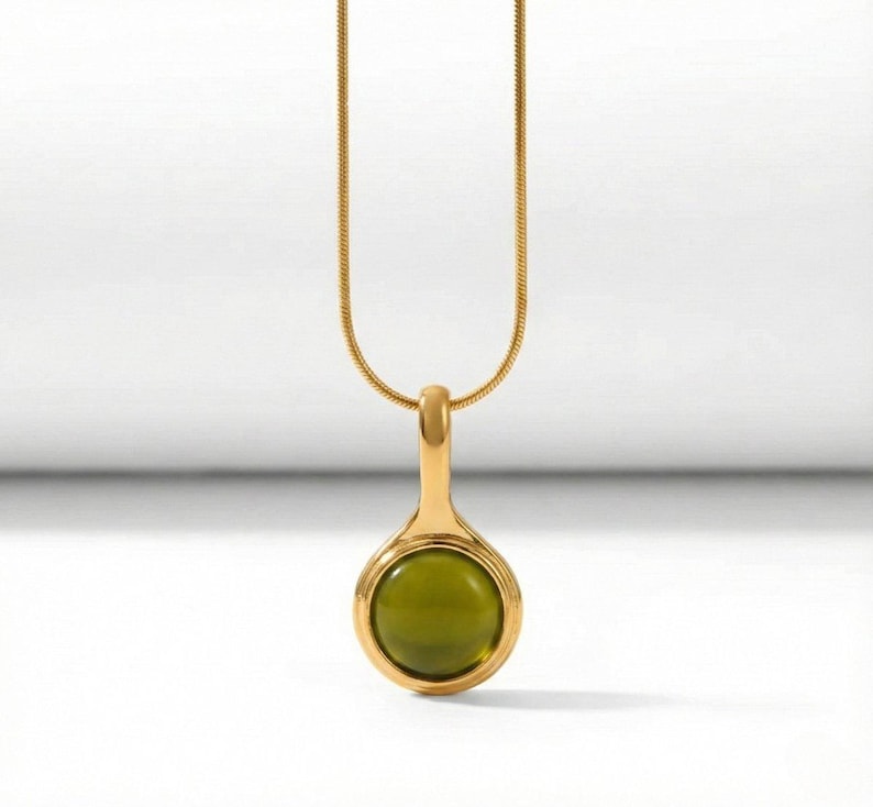 Op de afbeelding: Een goudkleurige ketting met een hanger met een ronde, olijfgroene steen. De hanger is gevat in een gouden zetting en hangt aan een fijne gouden ketting. De achtergrond is wit.