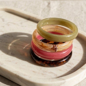 Op de afbeelding: Een stapel van vijf kleurrijke armbanden in groen, roze, bruin en bruin. De armbanden zijn gemaakt van een doorschijnend materiaal en zijn gestapeld op een wit marmeren dienblad. De armbanden zijn een stijlvol accessoire.
