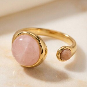 May include: A gold-colored ring featuring a large, round, light pink gemstone and a smaller, oval, light pink gemstone. The ring has an open design, with the gemstones set in a smooth, polished band.