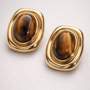May include: A pair of gold-coloured clip-on earrings. Each earring features a large, oval-shaped, brown and gold stone set within a gold frame. The earrings have a polished finish and a classic design.