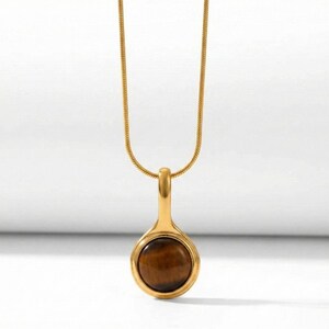 Op de afbeelding: Goudkleurige ketting met een hanger met een ronde, bruine tijgeroogsteen. De druppelvormige hanger hangt aan een fijne ketting. De ketting wordt getoond tegen een witte achtergrond.