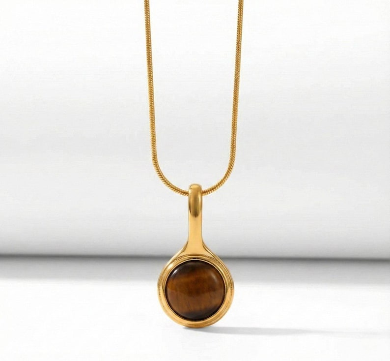 Op de afbeelding: Goudkleurige ketting met een hanger met een ronde, bruine tijgeroogsteen. De druppelvormige hanger hangt aan een fijne ketting. De ketting wordt getoond tegen een witte achtergrond.