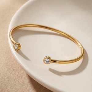 Pode incluir: Uma pulseira de punho aberta dourada com uma pedra preciosa redonda e transparente em cada extremidade. A pulseira está sobre um prato branco em forma oval. O fundo é de cor bege neutra.