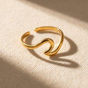 Può includere: Un anello color oro caratterizzato da un design fluido a forma di onda continua. La fascia è aperta nella parte superiore, creando una forma curva che ricorda l'aspetto di una singola cresta d'onda. La superficie metallica ha una finitura lucida.