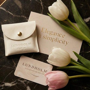 Op de afbeelding: Een cr&egrave;mekleurig su&egrave;de etui met een gouden drukknop, een kaart met de tekst "Elegance in simplicity" en een visitekaartje met de merknaam "Lundholm Scandinavian Design". Witte en roze tulpen zijn gerangschikt op een donker marmeren oppervlak.